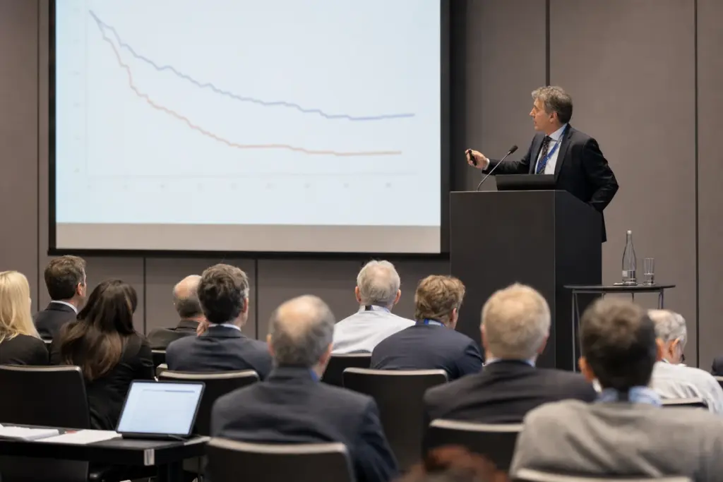 Relatore a un congresso medico davanti a una slide con grafici a linee, con il pubblico di professionisti seduto in sala