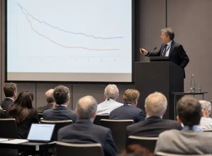 Relatore a un congresso medico davanti a una slide con grafici a linee, con il pubblico di professionisti seduto in sala