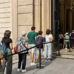 Coda di visitatori all’ingresso di un museo con barriera di sicurezza e portone aperto
