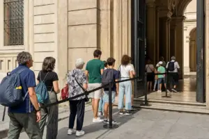 Coda di visitatori all’ingresso di un museo con barriera di sicurezza e portone aperto