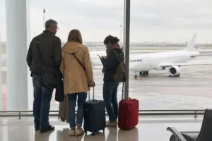 Tre viaggiatori con valigie aspettano al finestrone dell’aeroporto, con un aereo bianco sulla pista sullo sfondo