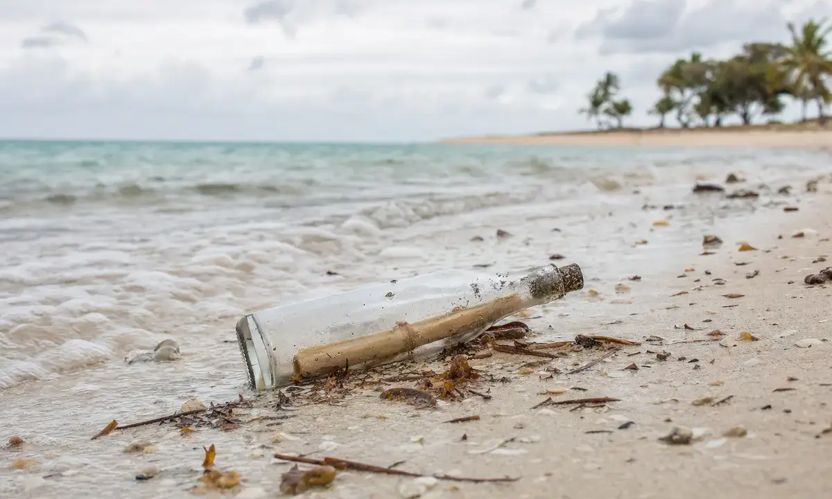 Bottiglia di vetro con un messaggio arrotolato sulla battigia di una spiaggia, con mare calmo sullo sfondo