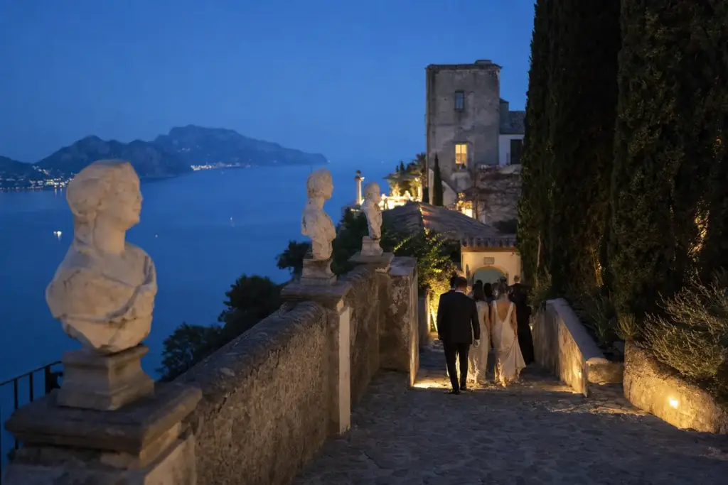 Ospiti eleganti scendono una scalinata in pietra tra busti in marmo con vista sul mare al tramonto