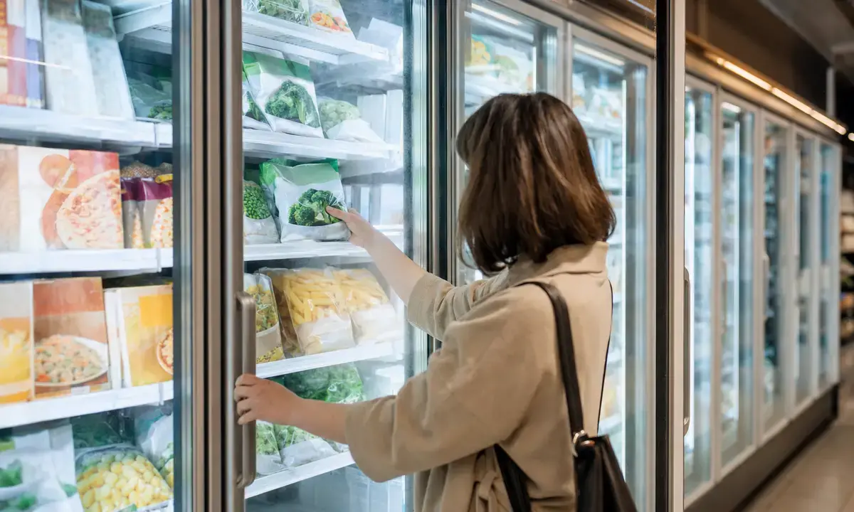 Cliente al supermercato apre un freezer a vetro e prende una busta di verdure surgelate
